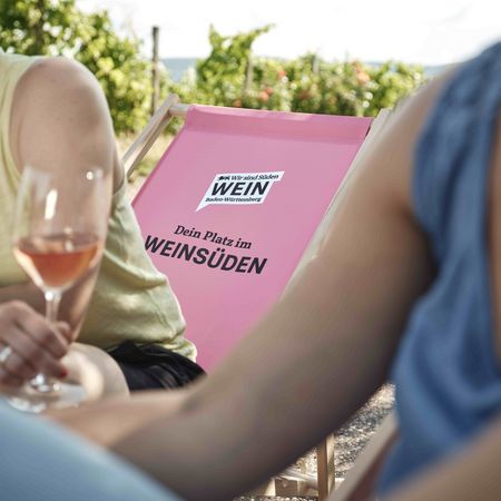 Dein Platz im Weinsüden: Genussvolle Momente im Land der 1000 Hügel Zwei Personen genießen Wein, im Fokus ein pinker Liegestuhl mit der Aufschrift ‚Dein Platz im Weinsüden‘ vor der Kulisse der Weinberge im Land der 1000 Hügel.