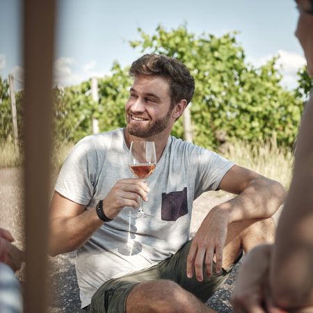 Entspannt genießen: Weinmomente im Kraichgau-Stromberg Ein junger Mann sitzt entspannt in den Weinbergen des Kraichgau-Stromberg, lächelnd mit einem Glas Roséwein in der Hand, während er sich mit Freunden unterhält.