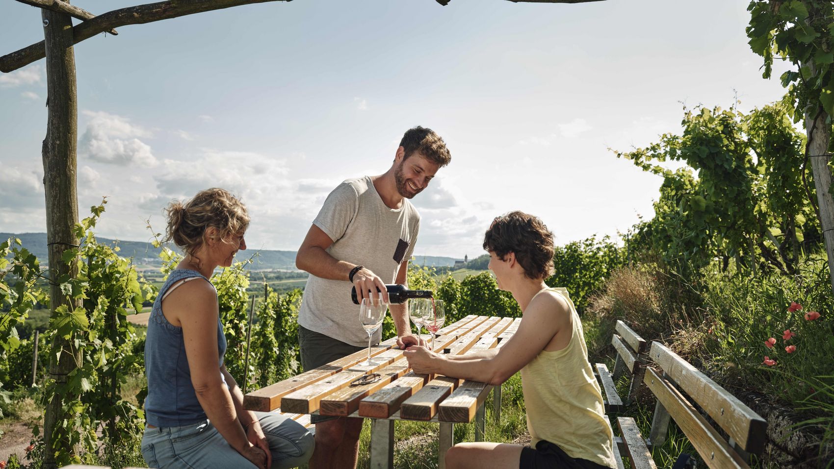 Weingenuss mit Aussicht: Weinprobe in den Weinbergen Brackenheims Drei Personen bei einer Weinprobe in den Weinbergen des Kraichgau-Stromberg, an einem rustikalen Holztisch unter einer Pergola sitzend, mit Blick auf die malerische Landschaft.