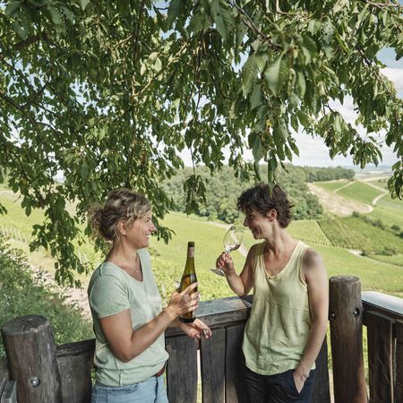 Weingenuss mit Aussicht: Unvergessliche Momente im Land der 1000 Hügel Zwei Frauen genießen Wein auf einem Aussichtspunkt im Land der 1000 Hügel, eine hält eine Weinflasche, die andere ein Glas Weißwein, umgeben von malerischen Weinbergen und Natur.