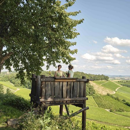 Aussichtsreiche Weinmomente: Genuss mit Panoramablick über Brackenheim Zwei Frauen genießen eine Weinprobe auf einem rustikalen Aussichtspunkt in den Weinbergen des Kraichgau-Stromberg, umgeben von grünen Rebhängen und einer malerischen Landschaft.