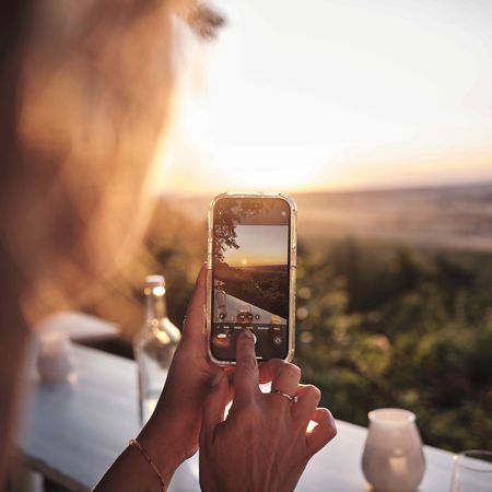 Sonnenuntergang & Weingenuss: Magische Momente im Land der 1000 Hügel Eine Person fotografiert einen Sonnenuntergang mit dem Smartphone, im Vordergrund ein gedeckter Tisch mit Wein und Kerzen, im Hintergrund die sanfte Landschaft des Land der 1000 Hügel.