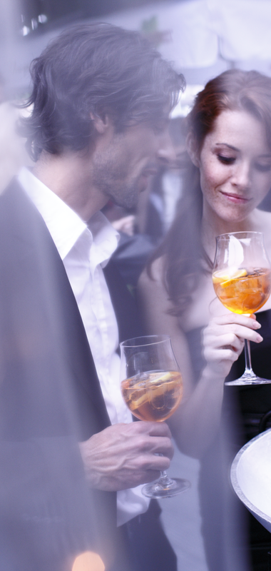 Ein elegantes Paar steht bei einer Veranstaltung im Freien, beide mit einem Glas Aperitif in der Hand. Die Frau, in einem schwarzen Abendkleid, betrachtet ihr Getränk mit einem sanften Lächeln, während der Mann in einem Anzug ihr nahe steht. Durch einen Schleier von Lichtern im Vordergrund wird eine romantische und stilvolle Atmosphäre geschaffen. Im Hintergrund sind unscharfe Silhouetten anderer Gäste und eine lebhafte Veranstaltung zu erkennen. | © TMBW | Christoph Düpper