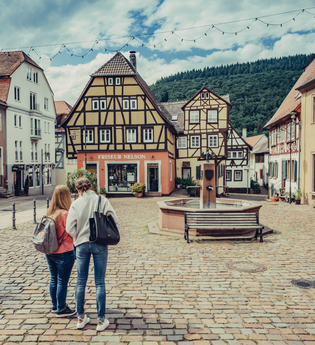 Ein Platz in einer Altstadt mit Kopsteinpflaster. In der Mitte ist ein Brunnen davor stehen zwei Frauen. Um den Platz stehen Fachwerkhäuser. Neckargemünd im Odenwald. | © Touristikgemeinschaft Odenwald e.V.