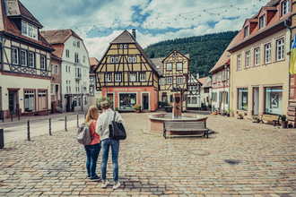 Ein Platz in einer Altstadt mit Kopsteinpflaster. In der Mitte ist ein Brunnen davor stehen zwei Frauen. Um den Platz stehen Fachwerkhäuser. Neckargemünd im Odenwald. | © Touristikgemeinschaft Odenwald e.V.