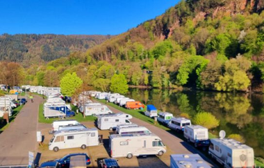 Campingplatz mit Wohnwägen und Wohnmobilen. Auf der rechten Seite ist ein Fluss (Neckar), dahinter am Ufer ein herbstlich bunter Wald. Neckargemünd im Odenwald