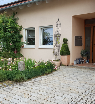 Eingangsbereich eines Hauses mit brauner Tür, Hausnummer 21, Blumenbeet mit Rosen und grünen Pflanzen neben gepflastertem Weg | © Familie Bokmeier