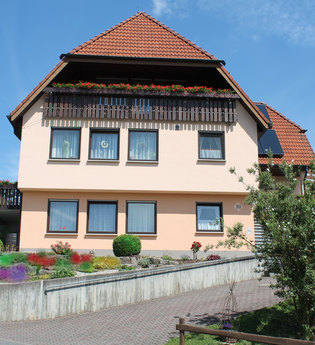 Zweistöckiges Ferienhaus mit roten Dachziegeln, Balkon mit Blumenkästen und gepflegtem Vorgarten unter blauem Himmel | © Ferienwohnung Ehrmann