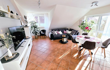 Helles Wohnzimmer mit braunem Fliesenboden, grauem Ecksofa mit Kissen, rundem Esstisch mit weißen Tischdecke und vier Stühlen, TV auf weißem Lowboard und großen Fenstern mit weißen Vorhängen | © Familie Fuhrmann