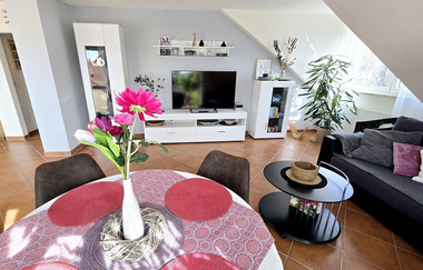Wohnzimmer mit rundem Esstisch, rosa Tischdecke und Blumen in Vase, weißem TV-Schrank, grauem Sofa mit Kissen und Pflanzen am Fenster | © Familie Fuhrmann