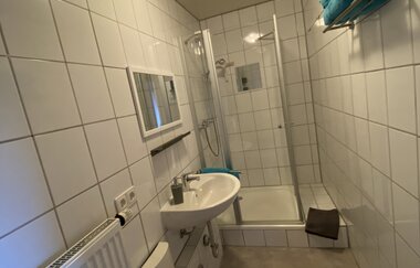 Modernes Badezimmer mit weiß gefliesten Wänden, Waschbecken, Spiegel, Duschkabine und Handtuchhalter mit blauen Handtüchern | © Ferienwohnung Etzl