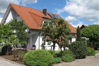 Weißes Einfamilienhaus mit rotem Ziegeldach und Solarpanelen, umgeben von grünen Büschen und blühendem Baum, bei sonnigem Himmel | © Erika Zeller