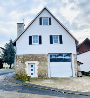 Zweistöckiges Haus mit weißer Fassade und steilem Dach, vier Fenstern mit schwarzen Fensterläden, weißer Haustür und großer Garagentür, im Hintergrund ein Glockenturm mit Uhr | © Ferienwohnung Etzl