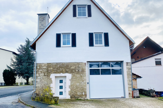 Zweistöckiges Haus mit weißer Fassade und steilem Dach, vier Fenstern mit schwarzen Fensterläden, weißer Haustür und großer Garagentür, im Hintergrund ein Glockenturm mit Uhr | © Ferienwohnung Etzl