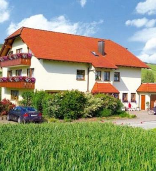 Zweistöckiges Haus mit rotem Dach, Balkonen mit Blumen, umgeben von grünen Feldern und zwei Personen mit Fahrrad vor dem Eingang | © Gästehaus Schieser