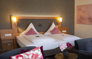 Einzelzimmer mit einem Bett, weißer Bettwäsche und orangefarbenem Kissen, Holzkopfteil mit zwei Wandlampen, rotem Teppich und Vorhängen neben einem Fenster | © Hotel Alexa