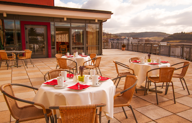 Terrasse eines Restaurants mit gedeckten Tischen, weißen Tischdecken, roten Servietten und Korbstühlen, im Hintergrund Hügel und bewölkter Himmel | © Savoy Hotel