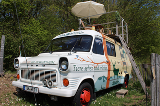 Ein umgebauter Bus zum übernachten. Eine Leiter führt auf der rechten Seite auf das Dach, wo sich eine kleine Terrasse mit Stühlen und Sonnenschirm befindet. | © Tourist-Information