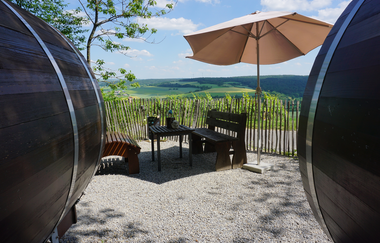 Eine Sitzgruppe steht vor den Weinfässern, der Bereich ist mit einem Sonnenschirm abgedeckt. | © Jakobshof Lehr