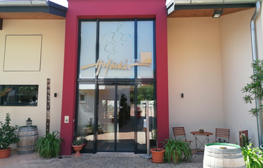 Blick vom Wohnmobilstellplatz auf das Eingangsportal mit großer Glasfront des Weinguts Hofäckers in Weikersheim-Queckbronn. Im Hof davor Sitzgruppe mit Pflanzen und Fässern als Deko. | © Touristinfo Weikersheim