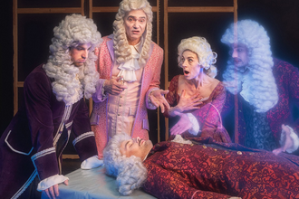 4 Personen mit Perücken stehen um einen Tisch auf dem eine weitere Person liegt