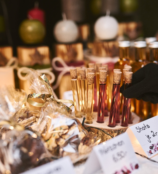 Produkte an einem Stand auf einem Weihnachtsmarkt (z.B. Shots und Gebäck). Eine Hand greift nach einem Shot im Reagenzglas. | © Touristikgemeinschaft Hohenlohe e.V. | Stefan Leitner Photography