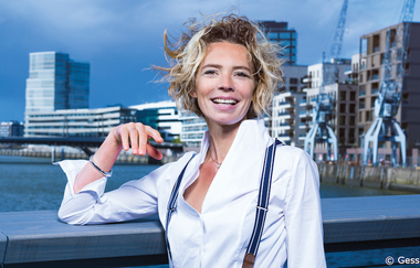 Frau mit lockigem Haar und weißem Hemd steht lächelnd vor einer urbanen Kulisse mit Wasser und modernen Gebäuden. | © Kultur- und Sportamt Stadt Bietigheim-Bissingen