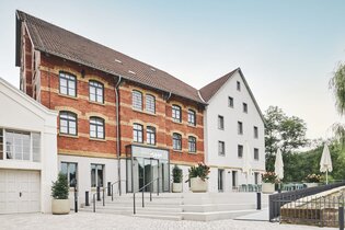 Zwei Gebäude, eines aus rotem Backstein und eines weiß, mit einem Eingang und Treppen. Blumenkübel und Tische mit Sonnenschirmen sind vorhanden. | © Kocherwerk