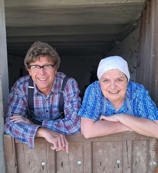 Zwei Personen lehnen sich lächelnd über eine Holztür, eine trägt ein kariertes Hemd, die andere ein blaues Kopftuch. | © SBEntertainment GmbH & Co. KG