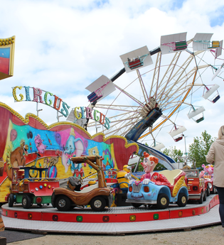 Auf dem Bild ist ein bunter Vergnügungspark mit einem Kinderkarussell, und einem Riesenrad im Hintergrund zu sehen. Mehrere Erwachsene und Kinder stehen davor oder gehen daran vorbei, während die farbenfrohen Fahrgeschäfte eine fröhliche Volksfestatmosphä | © Gemeinde Blaufelden