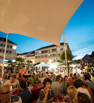 Gut mit Besuchern gefüllter Kiliansplatz in Abendstimmung. Die Kilianskirche ist beleuchtet und die Sonnensegel sind zu sehen.