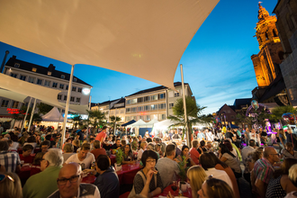 Gut mit Besuchern gefüllter Kiliansplatz in Abendstimmung. Die Kilianskirche ist beleuchtet und die Sonnensegel sind zu sehen.