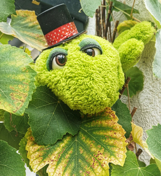 Ein grünes Plüschtier mit Zylinder schaut aus einem Blätterbusch hervor. | © Stadt Besigheim