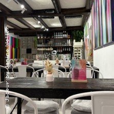 Ein stilvoll eingerichtetes Café mit modernen Kunstwerken an den Wänden und weißen Stühlen um dunkle Holztische. | © links im Bild