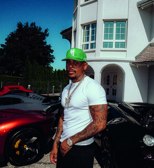 Luciano steht vor einem weißen Haus, neben sich zwei Autos.