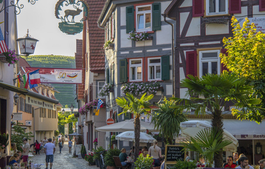 Eine malerische Fachwerkstraße mit Restaurants und Menschen, die spazieren gehen. Blumengeschmückte Häuser und Palmen säumen den Weg. | © Stadt Besigheim