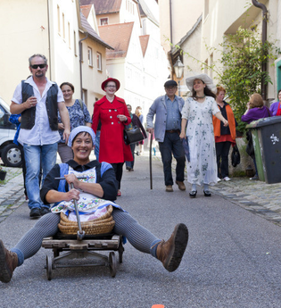 Eine Frau sitzt lachend auf einem kleinen Wagen in einer schmalen Gasse, umgeben von einer Gruppe fröhlicher Menschen. | © Stadt Besigheim