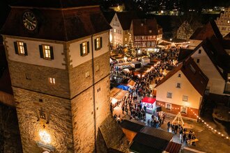 Nachtsicht eines beleuchteten Weihnachtsmarktes mit Menschen, die Stände besuchen, neben einem beleuchteten Turm mit Uhr.