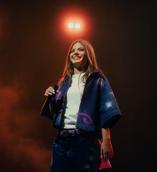 Die Künstlerin Zah1de steht auf der Bühne und hält ein Mikrofon in der Hand. Im Hintergrund leuchtet ein Scheinwerfer. | © William Krause