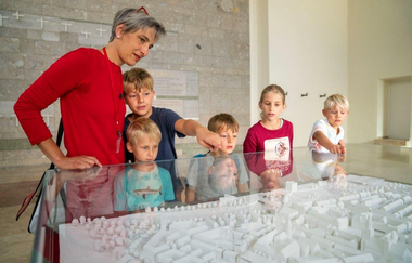 Das Bild zeigt eine Dame und fünf Kinder vor einem weißen Modell einer Stadt unter einem Glaskasten. Im Hintergund sowohl Stein- als auch beige Wände. Die Halle wirkt sehr geräumig.
