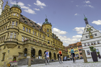 Blick auf den Marktplatz mit Rathaus in Rothenburg o.d.T.  Hier fahren gerade drei Radelnde über das Kopfsteinpflaster.