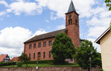 Kirchengebäude mit Kirchturm aus rotem Stein  auf einem mit Steinmauer umgrenzten Gelände und Bäumen | © Gemeinde Epfenbach