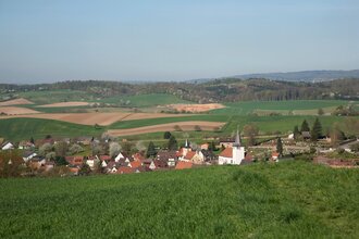 Blick auf das Dorf Spechbach mit mehreren Häusern und zwei Kirchen, umgeben von grünen Feldern und sanften Hügeln unter klarem Himmel. | © Landratsamt Rhein-Neckar-Kreis