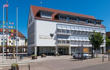 Weißes Verwaltungsgebäude mit heruntergelassenen Jalousien an einem gepflasterten Parkplatz | © Stadt Waibstadt