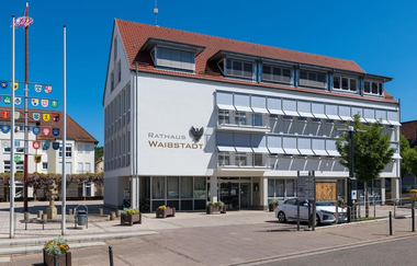 Weißes Verwaltungsgebäude mit heruntergelassenen Jalousien an einem gepflasterten Parkplatz | © Stadt Waibstadt