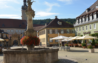Blick auf den Marktplatz in Weikersheim. Im Vordergrund ist ein großer Brunnen mit roten Geranien zu sehen. Dahinter Sitzmöglichkeiten mit Sonnenschirmen der Gastronomie. Im Hintergrund kann man das Schloss erkennen.