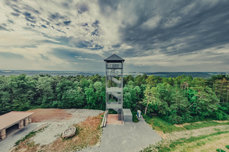 Luftaufnahme vom Aussichtsturm mit Nebengebäude auf dem Stahlberg in Külsheim-Uissigheim. Im Hintergrund geht der Blick ins bewaldete Taubertal.  Der Himmel zeigt ein wildes Wolkenschauspiel in dunkelblau und weiß. | © Liebliches Taubertal