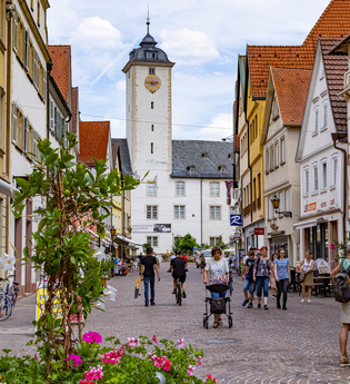 Blick in die Fußgängerzone in der Innenstadt von Bad Mergentheim. Es  sind viele Menschen unterwegs. Im Hintergrund ist das Residenzschloss und im Vordergrund Blumenkübel mit Sommerbepflanzung zu sehen.
