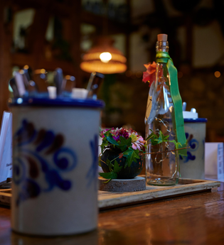 Tischdekoration mit Steingutkrügen, einer dekorierten Flasche und Blumenarrangement in einer Besenwirtschaft. | © Kraichgau-Stromberg Tourismus e.V.