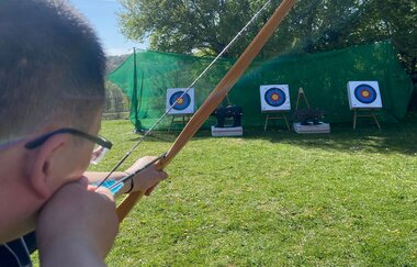 Ein Mann zielt beim Outdoor Bogenschießen mit einem Pfeil im gespannten Bogen auf eine Zielscheibe | © Touristikgemeinschaft Hohenlohe e.V. | Heffner Outdoor Events